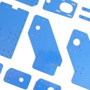 Puede incluir: Un surtido de placas metálicas de color azul brillante de varias formas y tamaños. Cada placa tiene múltiples agujeros perforados y algunas tienen recortes circulares más grandes. Las placas están dispersas sobre un fondo blanco.