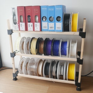 Può includere: Un'unità a scaffale in legno con tre ripiani contiene vari rotoli di filamento per la stampa 3D. I rotoli sono di diversi colori, tra cui bianco, nero, rosso, blu e verde. Gli scaffali sono etichettati con il marchio "Fiberlogy".