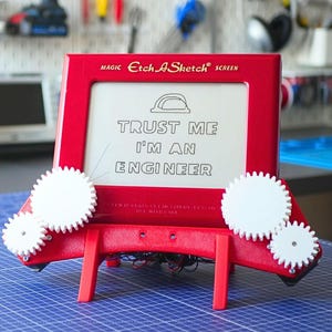 Peut inclure: Un écran Etch-A-Sketch rouge avec des engrenages blancs attachés. L'écran affiche le texte "TRUST ME I'M AN ENGINEER" avec un graphique de casque de chantier. Le produit est posé sur une surface quadrillée bleue.