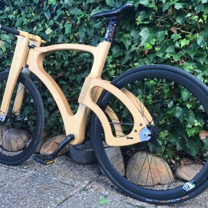 May include: A wooden bicycle with a unique frame design. The bike has black tires and a black seat. The frame is made of light-colored wood. The bike is parked on a stone surface in front of a green hedge.