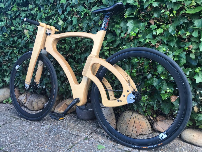 May include: A wooden bicycle with a unique frame design. The bike has black tires and a black seat. The frame is made of light-colored wood. The bike is parked on a stone surface in front of a green hedge.