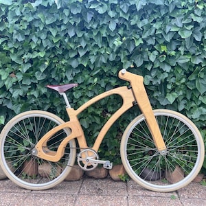 May include: A wooden bicycle with a unique frame design. The bike has a light brown frame, white tires, and a brown leather seat. The bicycle is set against a backdrop of green foliage and rocks.