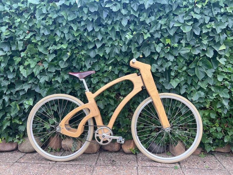 May include: A wooden bicycle with a unique frame design. The bike has a light brown frame, white tires, and a brown leather seat. The bicycle is set against a backdrop of green foliage and rocks.