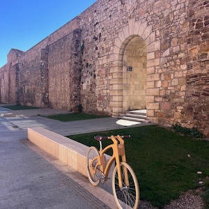 May include: A wooden bicycle with a light brown finish is parked on a concrete ledge. The bike has a unique frame design and white tires. The background features a large stone wall and a clear blue sky.