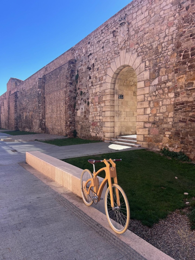 May include: A wooden bicycle with a light brown finish is parked on a concrete ledge. The bike has a unique frame design and white tires. The background features a large stone wall and a clear blue sky.