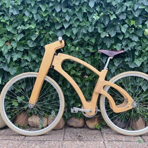 May include: A custom wooden bicycle with a unique frame design. The bike features light-colored wood, cream-colored tires, and a brown leather seat. The bicycle is set against a backdrop of green foliage and stone.