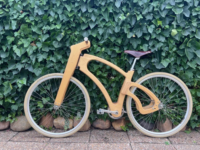 May include: A custom wooden bicycle with a unique frame design. The bike features light-colored wood, cream-colored tires, and a brown leather seat. The bicycle is set against a backdrop of green foliage and stone.