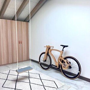 May include: A wooden bicycle with black tires and a black seat leans against a white wall. A gray swing hangs from the ceiling. A cream-colored rug with a black diamond pattern is on the floor.