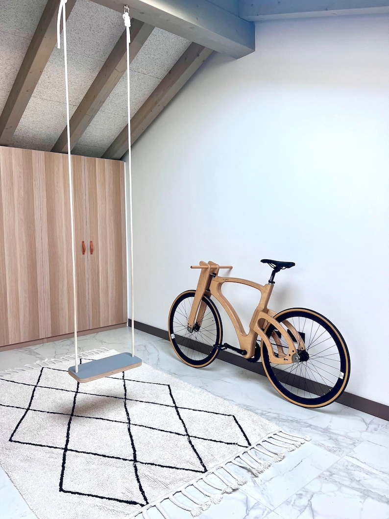 May include: A wooden bicycle with black tires and a black seat leans against a white wall. A gray swing hangs from the ceiling. A cream-colored rug with a black diamond pattern is on the floor.