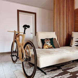 May include: A wooden bicycle with a unique frame design. The bike features black tires with a tan sidewall, a black seat, and a wooden handlebar. The bicycle is positioned in front of a white couch with decorative pillows and a patterned rug.