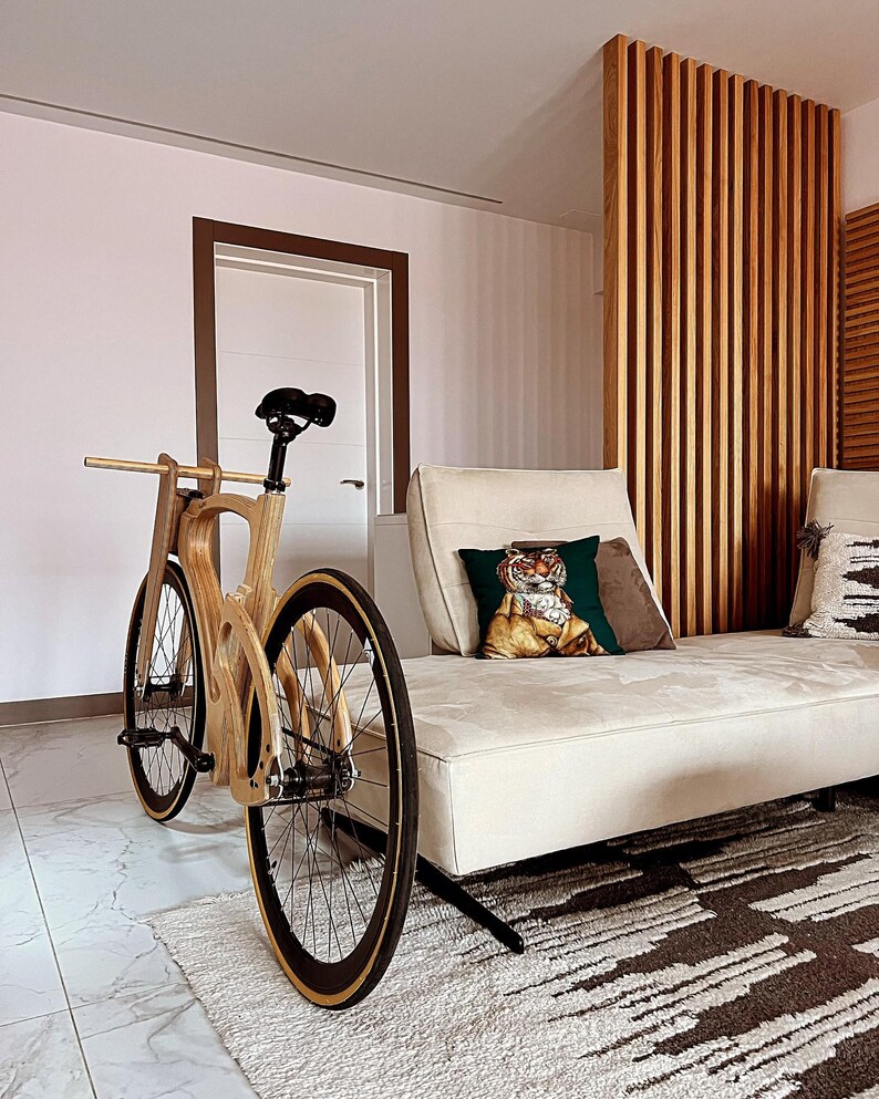 May include: A wooden bicycle with a unique frame design. The bike features black tires with a tan sidewall, a black seat, and a wooden handlebar. The bicycle is positioned in front of a white couch with decorative pillows and a patterned rug.