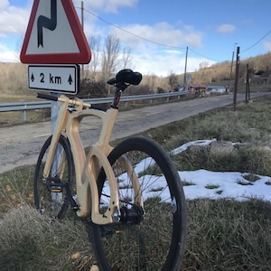 May include: A wooden bicycle with black tires and a black seat is parked on the side of a road. A road sign indicates a winding road ahead, 2 km away. The bike is made of light-colored wood and is set against a backdrop of a blue sky and sparse vegetation.