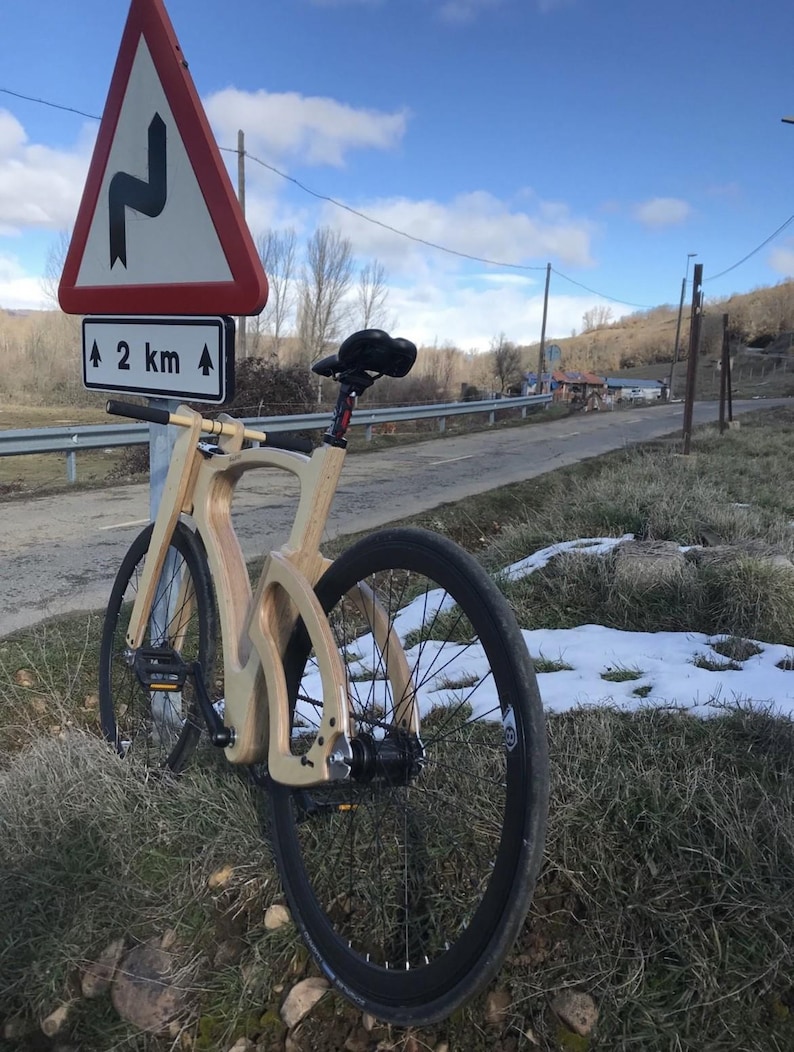 May include: A wooden bicycle with black tires and a black seat is parked on the side of a road. A road sign indicates a winding road ahead, 2 km away. The bike is made of light-colored wood and is set against a backdrop of a blue sky and sparse vegetation.