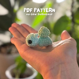 Op de afbeelding: Een kleine, gehaakte Bulbasaur-figuur in lichtblauw en groen, met een zwart oog. De tekst "PDF PATTERN no-sew bulbasaur" staat boven de figuur. De figuur wordt in een hand gehouden.