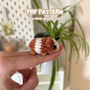 Puede incluir: Un pequeño peluche de cobaya hecho a mano y sin costuras. El peluche es marrón y blanco, con un aspecto suave y texturizado. El texto "PDF PATTERN no-sew guinea pig" es visible en el fondo. La cobaya mide aproximadamente 5 cm de largo.