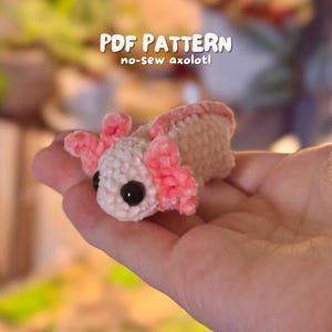 Puede incluir: Un ajolote de ganchillo en tonos crema y rosa se sostiene en una mano. El ajolote tiene branquias rosas y ojos negros. El texto "PDF PATTERN" y "no-sew axolotl" se muestra en letras blancas.