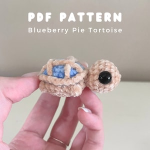 以下が含まれることがあります： 小さな編みぐるみのおもちゃで、亀の形をしています。亀は茶色で、青と白の甲羅をしています。おもちゃは大きな黒い目を持っています。