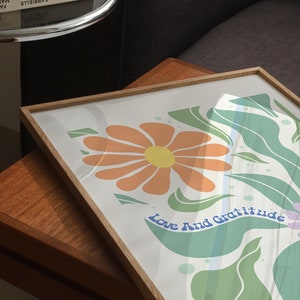May include: A framed print with a white background featuring a stylized orange flower with a yellow center and green leaves. The text "Love And Gratitude" is written in blue cursive letters.