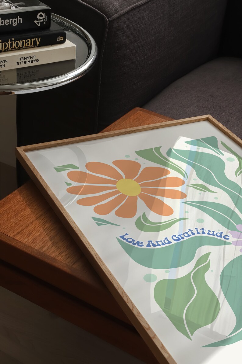 May include: A framed print with a white background featuring a stylized orange flower with a yellow center and green leaves. The text "Love And Gratitude" is written in blue cursive letters.