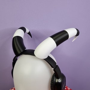Puede incluir: Auriculares rayados en blanco y negro con diadema blanca y auriculares negros. Los auriculares tienen un diseño único con dos cuernos blancos y negros en la parte superior.