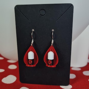 Può includere: Un paio di orecchini rossi e neri con un design cilindrico bianco. Gli orecchini sono a forma di goccia e pendono da ganci d'argento. Gli orecchini sono esposti su una carta nera.