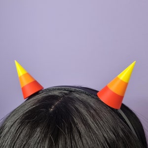 Puede incluir: Una diadema negra con dos cuernos en forma de maíz dulce naranja, amarillo y rojo unidos a ella.