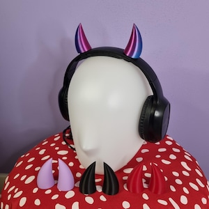Puede incluir: Un auricular inalámbrico negro con una cabeza de maniquí blanca. El auricular tiene dos cuernos de diablo rosas y azules unidos a la parte superior. Hay cuatro cuernos de diablo adicionales en la imagen, dos rosas, uno negro y uno azul.