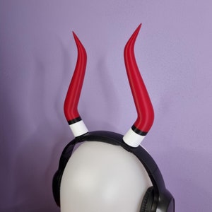 May include: Red devil horns attached to a black headband, with white and black accents. The horns are curved and pointed, and the headband is worn on a white mannequin head. Black headphones are also on the mannequin head.