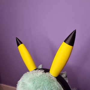 May include: A pair of yellow and black cosplay ears, resembling a popular cartoon character, attached to a black headband. The ears are bright yellow with black tips, set against a soft, light green plush background.