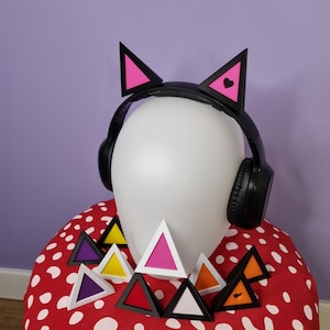 Puede incluir: Auriculares negros con orejas de gato rosas y un pequeño corazón rosa en cada oreja. Los auriculares están sobre una superficie de lunares rojos y blancos con una variedad de objetos triangulares de colores.