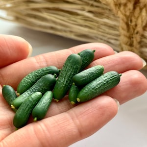 Könnte beinhalten: Eine Ansammlung von zehn Miniatur-Gurken in Dunkelgrün mit unebener, strukturierter Schale und kleinen gelben Spitzen an den Enden. Diese winzigen, realistischen Gemüsereplikate sind wie herkömmliche Gurken geformt und durchgehend dunkelgrün.