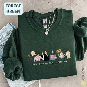May include: Forest green sweatshirt featuring the phrase "WHAT EXCELLENT BOILED POTATOES" and a graphic of figures, books, and a teapot. A silver necklace with a pendant is on the sweatshirt.