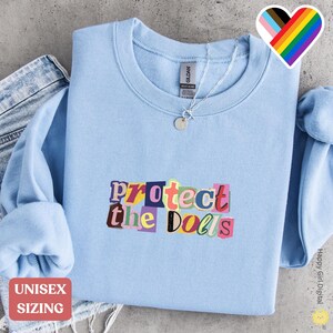 May include: A light blue unisex sweatshirt with the phrase "protect the Dolls" in colorful, cut-out style letters. The sweatshirt has a crew neck and long sleeves. A silver necklace is visible. The words "Unisex Sizing" are in a red box.