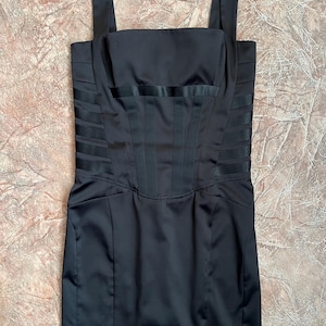 Puede incluir: Vestido negro de satén con escote cuadrado y tirantes anchos. El corpiño presenta detalles de pliegues verticales y horizontales, creando una silueta estructurada. El vestido tiene cintura entallada y falda recta.