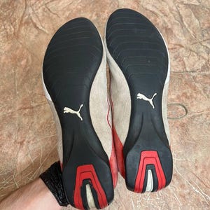 May include: Pair of Puma sneakers with beige suede uppers, red accents, and black soles. The soles feature a white Puma logo and red and white detailing. The shoes are designed for athletic use.