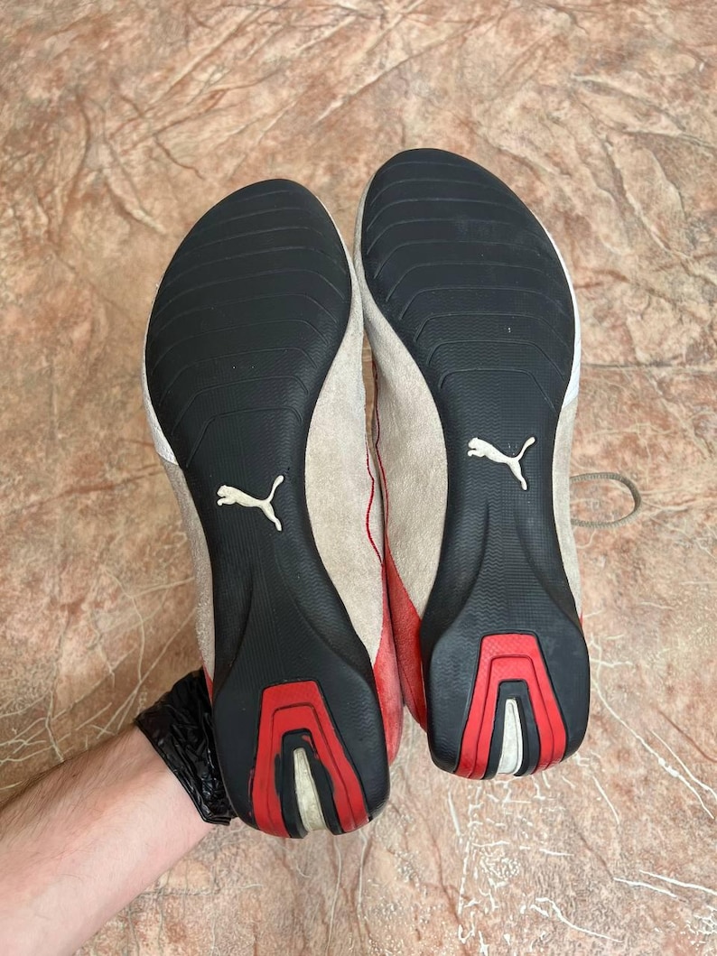 May include: Pair of Puma sneakers with beige suede uppers, red accents, and black soles. The soles feature a white Puma logo and red and white detailing. The shoes are designed for athletic use.