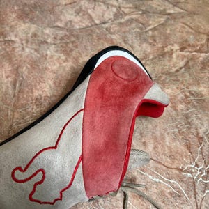 May include: A close-up of a gray and red shoe with a bird-like design. The shoe features a red suede panel on the front, a gray suede body, and a black and white trim. Red stitching outlines a flame-like design on the side.