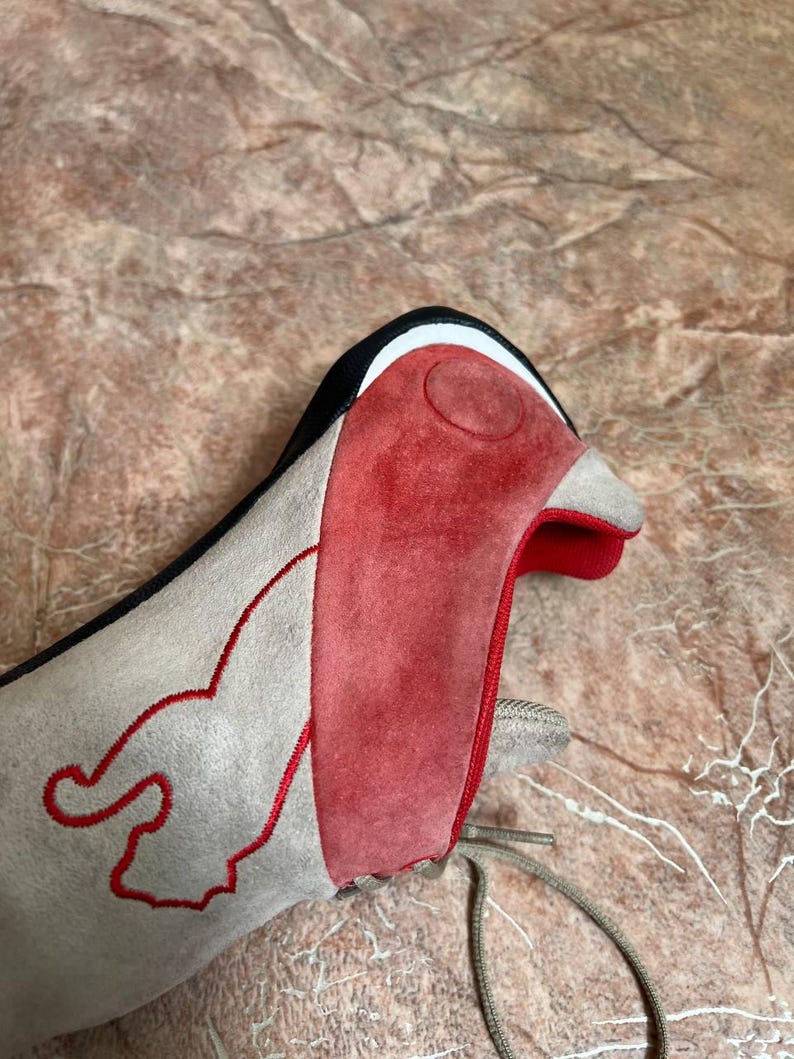 May include: A close-up of a gray and red shoe with a bird-like design. The shoe features a red suede panel on the front, a gray suede body, and a black and white trim. Red stitching outlines a flame-like design on the side.