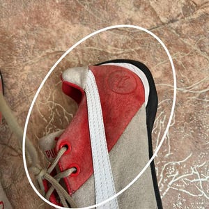 May include: Close-up of a Puma sneaker. The shoe features a red suede toe and heel, with a white stripe along the side. The laces are beige, and the eyelets are red. The shoe is on a brown surface.