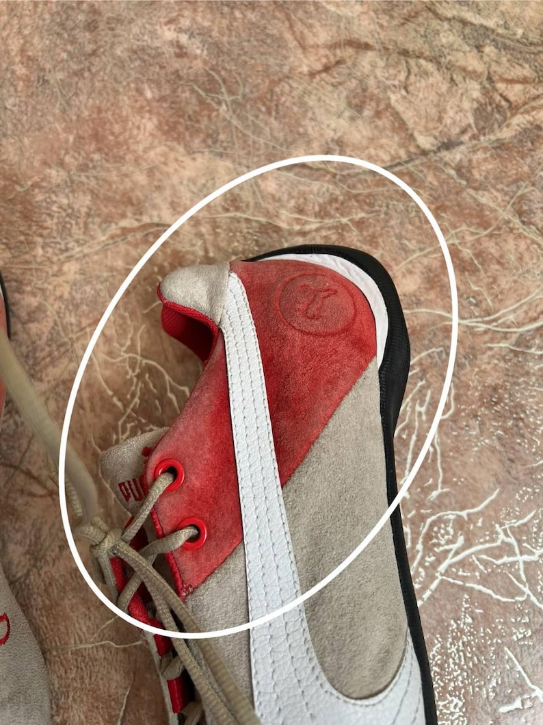 May include: Close-up of a Puma sneaker. The shoe features a red suede toe and heel, with a white stripe along the side. The laces are beige, and the eyelets are red. The shoe is on a brown surface.