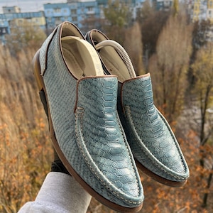 Może przedstawiać: Para niebieskich skórzanych mokasynów ze wzorem skóry węża. Buty mają brązowe wykończenia i podeszwy, z białymi detalami szwów. Mokasyny są podtrzymywane przez prawidło do butów.