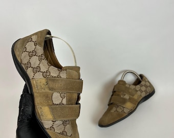 May include: Tan suede trainers featuring the Gucci logo pattern. The trainers have two hook-and-loop straps and a black sole. The shoes are displayed on a white background.
