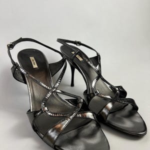 May include: A pair of black high-heeled sandals with a metallic sheen. The sandals feature thin straps, a crisscross design over the foot, and a slender heel. The brand name is visible on the insole.