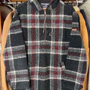May include: A black, white, and red checked hooded jacket with a half-zip and drawstring. The jacket features a small logo patch and is suspended on a wooden hanger.