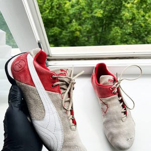 May include: Pair of Puma sneakers in beige suede with red accents. Features a white Puma logo on the side and red detailing around the ankle and tongue. The sneakers have beige laces and a black sole.