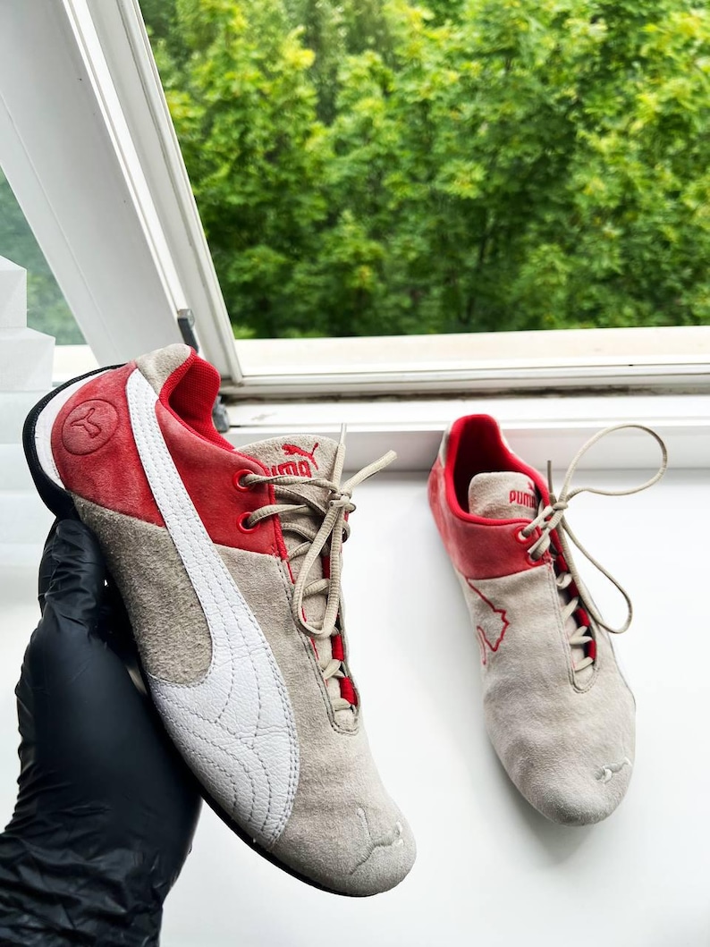 May include: Pair of Puma sneakers in beige suede with red accents. Features a white Puma logo on the side and red detailing around the ankle and tongue. The sneakers have beige laces and a black sole.