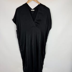 May include: A solid black midi-length dress with a deep V-neckline and short, wide sleeves. The garment features a smooth, slightly reflective fabric finish and a relaxed, loose-fitting silhouette that hangs straight from the shoulders.