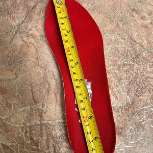 May include: A red shoe insole is measured with a yellow tape measure. The insole is approximately 10 inches long. The insole is red with a white logo. A portion of a red and black shoe is visible in the upper left corner.