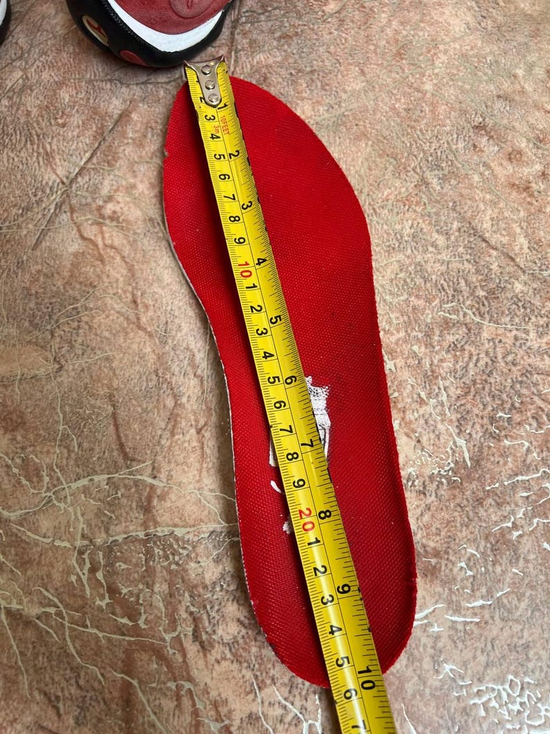 May include: A red shoe insole is measured with a yellow tape measure. The insole is approximately 10 inches long. The insole is red with a white logo. A portion of a red and black shoe is visible in the upper left corner.