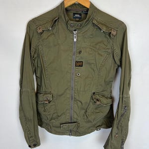 May include: Olive green jacket with multiple pockets, a zip-up front, and a small logo patch. The jacket has a stand-up collar and is hanging on a wooden hanger. The jacket is made of a durable material.